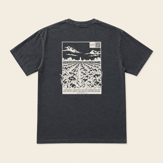 A dark gray crewneck t-shirt laid flat on an ivory background, showing a large white graphic print on the back depicting a cotton field under a cloudy sky.